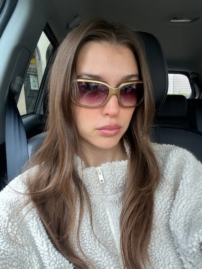 A young female is depicted seated in the front passenger seat of a vehicle in Loire-Authion, France. She has long brown hair, parted centrally, and is wearing a cream-colored, textured quarter-zip fleece pullover with a white zipper. Her eyes are obscured by large, rectangular sunglasses featuring gold-toned frames and gradient purple-tinted lenses. Her lips are closed and coated with a light pink gloss. A black vehicle seatbelt is positioned across her left shoulder and chest. Through the vehicle's left-side window, a blurry exterior shows a building with a yellow commercial sign, possibly from a LIDL supermarket, displaying indistinct text and partial numbers (e.g., '3.90'). A black vehicle headrest is visible behind her, and blurred green foliage is discernible through the rear passenger window.