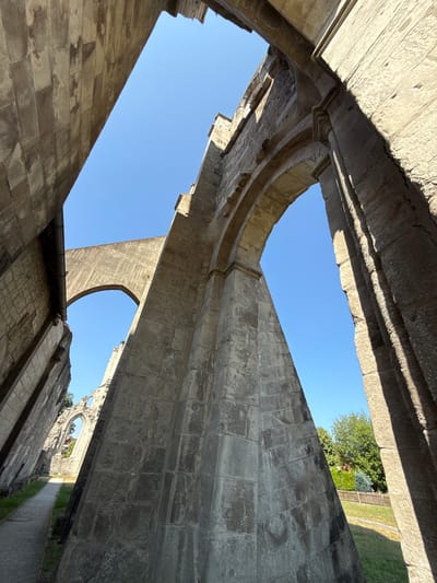 The image captures the ruins of the Walkenried Abbey in Walkenried, Germany. The viewpoint is looking upwards through massive stone arches and walls, showcasing the architectural remnants of the historical abbey. The sky is a clear, bright blue. The stone structures are weathered and show signs of age, with visible masonry and varying textures. In the distance, glimpses of green trees, a wooden fence, and some modern buildings are visible, indicating the abbey is situated within or adjacent to a more contemporary environment. A paved pathway can be seen leading through the ruins. There are no people present in the image.