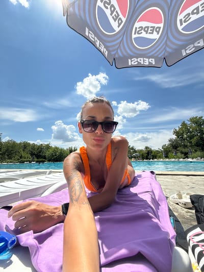 A woman wearing sunglasses and an orange bikini is lying on a purple towel on a lounge chair next to a swimming pool. She has tattoos on her arm. A Pepsi umbrella is overhead. The pool area is surrounded by trees and lounge chairs. The sky is blue with white clouds. The location is Grivitsa, Bulgaria.