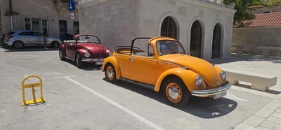 Two classic Volkswagen Beetle convertibles, one maroon and one bright orange, are parked side-by-side on a paved surface in what appears to be a town square or parking area in Općina Pučišća, Croatia. The maroon Beetle is parked behind the orange one, slightly angled to the left. The orange Beetle is prominently positioned in the foreground, facing to the right, with its top down. A parking barrier, painted bright yellow, is visible in the lower left quadrant of the image. In the background, a stone building with arched entrances and a tiled roof is visible, suggesting a Mediterranean architectural style. A silver hatchback car is parked further back, to the left of the Beetles, with a person standing beside it. The ground is paved with concrete or asphalt, and white parking lines are marked on the surface. The scene is bathed in bright sunlight, casting shadows from the vehicles and a bench in the background. The overall impression is a sunny day in a picturesque European town.