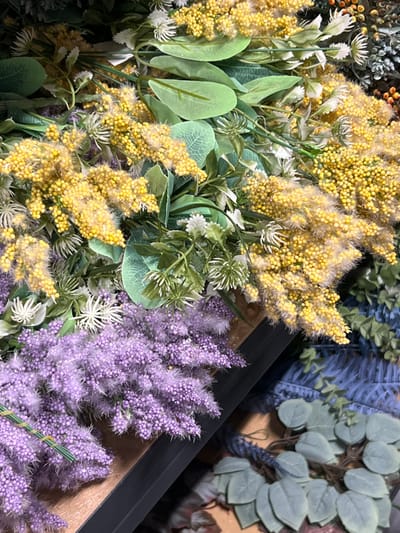 The image displays a close-up, static scene of various artificial floral arrangements, likely in a retail or decorative display setting in Warzymice, Poland. The foreground and mid-ground are dominated by bundles of yellow, mimosa-like artificial flowers with a fluffy texture, intermixed with light green synthetic leaves and occasional small white spiky artificial blossoms. Below these, and extending towards the lower left, are several bundles of light purple, lavender-like artificial flowers, characterized by a fine, bristly texture, some secured with green floral wire. Towards the right and in the background, additional artificial foliage is visible, including blue, fern-like leaves and bundles of grayish-green, rounded, eucalyptus-like artificial leaves. The items are arranged on what appears to be a dark-colored shelf or display surface. No individuals or active interactions are present in the scene.