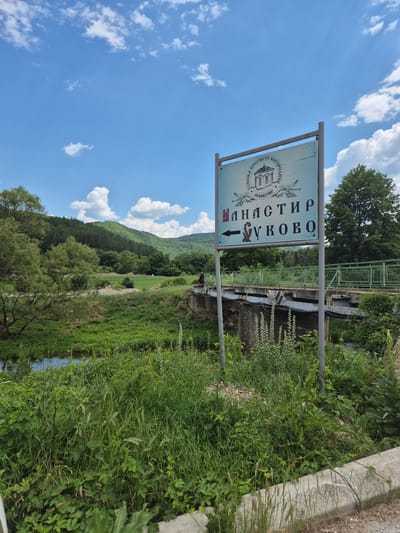 The image shows a sign for "Manastir Sukovo" pointing left, indicating a monastery. The sign is in Bulgarian script. It stands in front of a bridge that spans a small body of water, likely a river or stream. The area is lush and green, with trees and hills in the background. The sky is blue with scattered white clouds. A person is visible on the bridge. The environment appears to be rural.