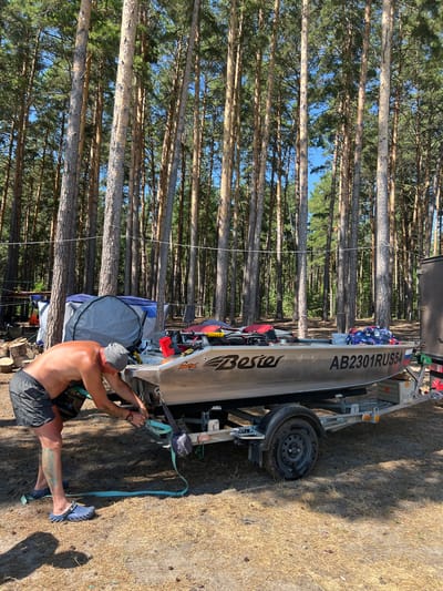 A man is seen securing a boat to a trailer in a forested area. The boat, which has "Bester" and "AB2301RUS54" written on its side, is silver and rests on a gray trailer. The man, wearing dark shorts and a grey beanie, is leaning over and appears to be fastening a strap to the boat's engine. A blue tent is visible in the background, along with stacks of firewood and other camping equipment. The ground is covered in dry grass and pine needles, and tall pine trees surround the scene. The sky is clear and blue. The location is the city of Borovoye, Russia.