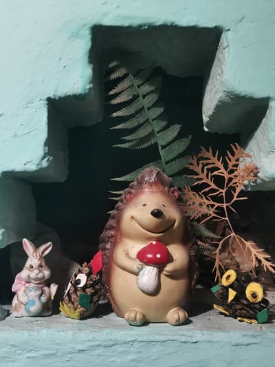 A whimsical display sits within a pale blue, textured niche. A plump hedgehog figure, clutching a red-and-white mushroom, anchors the arrangement. To its left, a ceramic rabbit with a blue jar and a pinecone owl with googly eyes add charm. To the hedgehog's right is another pinecone owl. A fern frond and a brown, dried branch provide a natural backdrop, enhancing the scene's playful, woodland aesthetic.