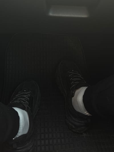 A top-down indoor shot depicts a pair of black athletic shoes being worn with white socks and black pants in the footwell of a vehicle. The footwell is lined with a black rubber mat with a square grid pattern. To the left is a smooth, dark gray or black surface, possibly the car's interior. The location is unknown, and there is no mention of Diadovo, Bulgaria, in the image.