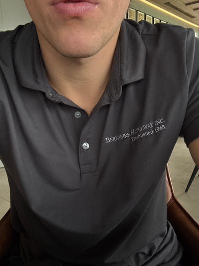 The image shows a person wearing a dark grey polo shirt with "BERKSHIRE HATHAWAY INC. Established 1965" embroidered on the upper left chest. The shirt has visible buttons down the front. The person is facing the camera, with only part of their face, including lips and neck, visible. The background shows a hint of a tiled floor and the edge of a chair. The overall perspective is slightly tilted.