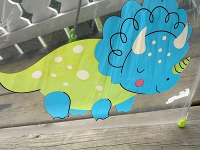 A stylized, cartoonish blue triceratops with a lime green body is depicted on a transparent surface, likely plastic, which is resting on a weathered wooden plank surface. The dinosaur has white horns, a curly mane detail on its head, a pink cheek, and a small black eye. The green body is adorned with several white polka dots and transitions into blue legs and a tail. The background is out of focus, showing a metallic grate-like structure above and what appears to be greenery. The image was taken in Ridabu, Norway.