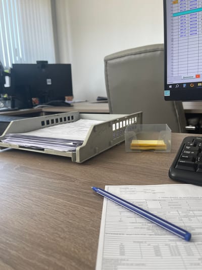The image shows a close-up view of a wooden desk with various office supplies. In the foreground, a blue pen lies diagonally across a document. To the left, a gray document tray is filled with papers. Behind it, a small clear plastic holder contains yellow sticky notes. In the background, a computer monitor displays a spreadsheet with Cyrillic text, and a portion of a gray office chair is visible. The environment appears to be an office setting, though no specific location in Pyt'-Yakh, Russia, can be determined from the visual information provided.