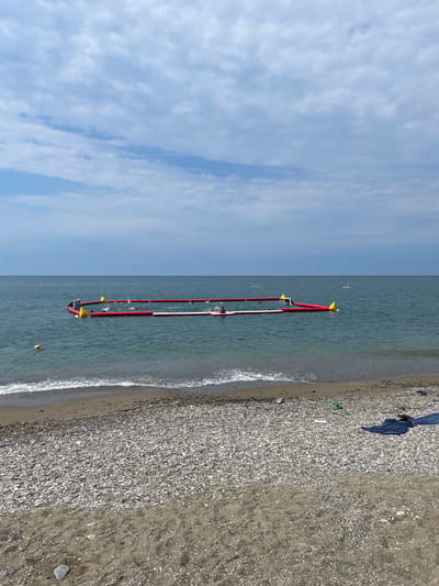 A wide shot captures a section of the Black Sea coastline in Krinitsa, Russia. In the foreground, a pebble beach transitions into dark sand. Waves gently lap the shore. A red and white inflatable structure, appearing to be a water sports arena, floats in the turquoise water. Several yellow buoys are positioned around it. A few people are visible on the inflatable structure, and some individuals are on paddleboards further out in the sea. A small beach towel and a pair of green flip-flops are on the shore. The sky is partly cloudy with soft grey and white clouds.