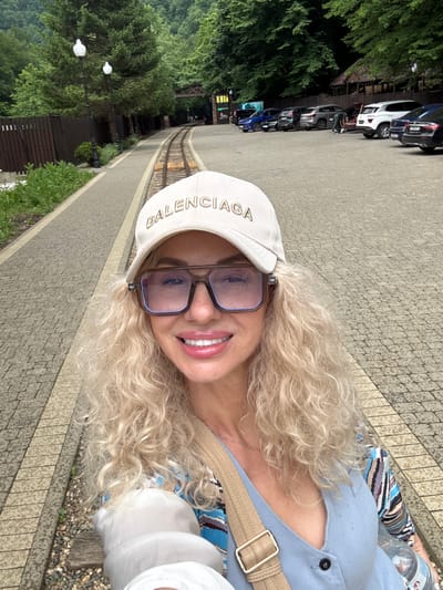 A woman with curly blonde hair, wearing sunglasses, a light blue shirt, and a cream-colored baseball cap with "BALENCIAGA" printed on it, smiles at the camera. She is outdoors, standing on a paved walkway with a stone pattern. Behind her, a row of cars is parked along a road, and to her left, lush green trees and foliage create a natural backdrop. The scene appears to be set in a park or a scenic area. The lighting suggests it is daytime, possibly overcast as there are no strong shadows. The woman's expression conveys happiness. In the background, there is a glimpse of what looks like a small train track.