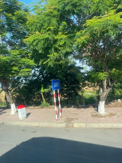 The image depicts a roadside scene in Diên Lạc, Vietnam, with lush green trees dominating the background and foreground. A blue sign with "AH1" in white letters is prominently displayed, supported by a red and white striped pole. To the left, a concrete marker with a red top displays "Q: 0" and "38Km". The ground is paved with reddish-brown bricks, and a sidewalk separates it from the asphalt road in the foreground. The area is well-lit, suggesting it is daytime. The environment appears to be a relatively quiet, possibly rural or suburban, area.