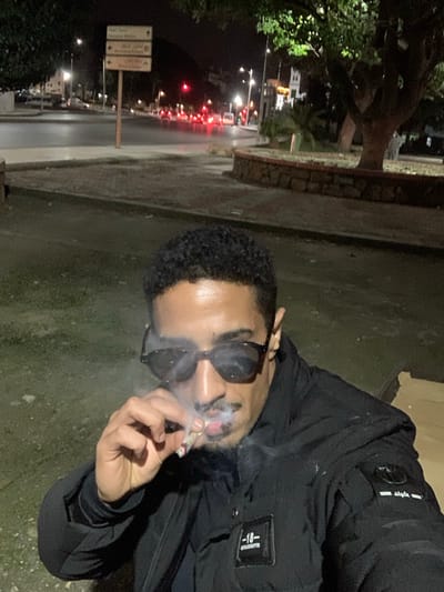 A man with short, dark hair and sunglasses smokes a cigarette in the dark. He's wearing a black jacket with visible labels. In the background is a dimly lit street at night, with trees, streetlights, buildings, and red taillights indicating traffic. The image is slightly blurry and appears to be taken at a low angle.