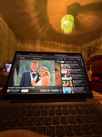 A laptop is on a table in a room. The screen shows a YouTube video titled "Четирима студенти, една жестока съдба" (Four students, a cruel fate) by the channel "Guilty as Charged." The video thumbnail shows a smiling couple. In the background, there is a lamp with a decorative green lampshade, and a TV showing some kind of broadcast. The room appears to be a bedroom or living room, but the location in the city of Dobrovnitsa, Bulgaria, cannot be verified from this image. There is no discernible interaction between people.