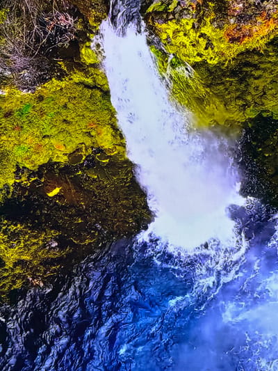 The media file depicts an aerial view of a waterfall cascading into a dark, turbulent body of water. The surrounding landscape is characterized by lush, moss-covered rocks and vegetation, with some areas showing a vibrant yellow-green hue, while others appear darker and more muted. The waterfall itself is a frothy white, creating a stark contrast against the deep blue of the water and the verdant rocks. The water's surface is textured with ripples and waves, suggesting movement and a natural, wild environment.

The scene appears to be set during daylight, though the specific time of day is not discernible. The weather appears to be clear, with no visible atmospheric conditions like rain or fog. There are no people or man-made objects in the frame, emphasizing the natural beauty of the landscape. The perspective is from above, giving a sense of scale and the grandeur of the waterfall.

Notable details include the intense color saturation of the moss and rocks, which could be indicative of moist conditions. The dynamic movement of the water, both in the cascade and the pool below, is a prominent feature. The overall impression is one of raw, untamed nature. Given the location context of Gorzow, Poland, this could be a natural landmark or a carefully preserved natural area. No text is visible in the media file.