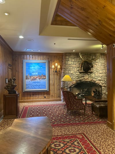 The image showcases an interior with rustic design elements. A stone fireplace with a mounted moose head sits atop a wall. Below, a painting hangs above a wooden console table with decorative sculpture. A leopard-print chair rests near the fireplace. A patterned rug in red and beige covers the floor, leading to a wooden table. The walls are wood paneling, and the ceiling is slanted, with wood planks and recessed lighting.