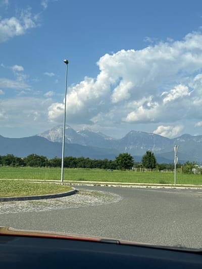 The photograph depicts a road in the city of Kranj, Slovenia, with the Alps visible in the background. A grassy median runs alongside the road, dotted with trees and streetlights. The sky is blue with scattered white clouds. A portion of a vehicle is visible on the right edge of the frame.