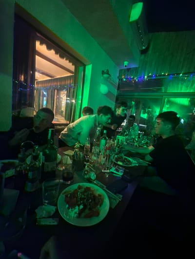A medium indoor shot shows a crowded table setting, presumably in a restaurant or bar. Several people are seated around the table, seemingly engaged in a meal or social gathering. The lighting has a strong green hue, casting a distinctive tone over the scene. A plate of food, possibly a salad, is prominently placed on the table. Numerous empty bottles and glassware are scattered around, indicating consumption. A window with decorations and a partial view of an illuminated exterior suggests that this location might be in the city of Panagyurski kolonii, Bulgaria.
