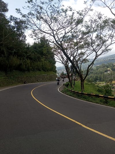 This image displays a curved asphalt road on a hillside in Gubbisamamidi, India. The road is marked with a solid yellow center line and gently curves left into the mid-distance. To the immediate left, a textured stone retaining wall supports a steep embankment covered in dense, dark green foliage and trees. Along the right side of the road, a black and yellow metal guardrail runs parallel to the curve, overlooking a downward slope. Several trees with prominent, bare branches and sparse green leaves are positioned between the road and the guardrail. In the mid-ground, three motorcycles, each with a rider, are observed on the road, navigating the curve, moving away from the viewer. Beyond the guardrail and trees on the right, a wide panoramic view reveals a valley containing scattered buildings and structures amidst abundant green vegetation, extending towards distant hills under a partly cloudy sky. Overhead power lines are visible crossing the valley in the background.