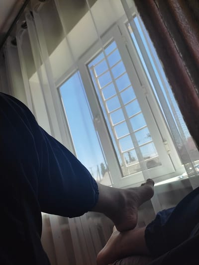 The image shows a close-up view of a person's legs and feet in the foreground, with a window and curtains in the background. The person is wearing dark blue pants. The window is open, revealing a bright blue sky and a white window frame with a grid pattern. Light, sheer curtains hang on either side of the window. The right side of the image also shows a darker, textured curtain. The scene appears to be indoors, likely in a residential setting. The location is specified as Ала-Арча, Kyrgyzstan.