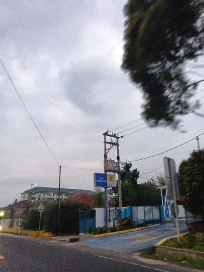 A street scene captured under an overcast sky features a utility pole equipped with a transformer and multiple power lines on the right side of a paved road. Positioned below the utility pole is a blue rectangular sign displaying the text "PLN CIMACAN" and "PLN" with an associated logo. This signage identifies the location as being in Indonesia, specifically Cianjur/Cimacan, which contradicts the specified Phước Hòa, Vietnam.
The foreground presents a paved road, displaying a faint yellow line on its left edge, leading towards a blue-paved ramp. This ramp provides access to a gated facility, secured by a light blue fence. Dense green foliage and trees are visible surrounding the gated compound and along the roadside. In the background, a multi-story building with a light green roof and numerous windows is discernible above the tree line. A single artificial light source is illuminated in the far-left background. The upper right portion of the image shows a significantly blurred section of a tree, indicative of motion during the photograph's capture. No individuals or vehicles are present in the scene. The overall lighting is diffused, consistent with an overcast day or twilight conditions.