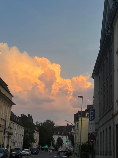 This is an eye-level, medium shot of a street in Herne, Germany, at sunset. The sky is filled with large, luminous clouds, painted in shades of orange and yellow by the setting sun. They are dramatic and fill much of the upper frame.
On the right side of the image is a multi-story building with a grey facade, featuring rows of windows and decorative architectural elements. A street lamp is visible in front of it. A blue parking sign with an arrow pointing left, indicating a parking area, is mounted on a pole. Below it, a circular sign with a red border and a diagonal red bar through a blue circle indicates "no stopping."
On the left side of the street are several buildings. The closest building on the left has a yellow facade with classical architectural details and a dark tiled roof. It has a blue flag with a crest hanging from a balcony. Further down the street, more buildings with dark roofs and light-colored facades are visible, interspersed with trees.
Several cars are parked and driving on the street. In the foreground on the left, a dark-colored van or SUV is parked. Further down, a black Audi, a white sedan, and a dark SUV are visible. Traffic lights show a green signal. The street appears to be paved, with painted lane markers. The overall atmosphere is serene, with the dramatic sunset clouds dominating the scene.
