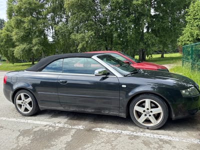 A black Audi A4 convertible is parked on an asphalt surface next to a white line. The car's top is down, revealing a tan interior. The car is positioned sideways in the frame, and the image is rotated 90 degrees counterclockwise. In the background, a red convertible is partially visible, also with its top down. Beyond the cars, there is lush green grass, trees, and a green metal fence. The sky appears overcast, suggesting it might be a cloudy day. The overall scene depicts a parking area near a natural, green setting. No people are visible in the image. There is no visible text in the image. The location context is Haabneeme alevik, Estonia.