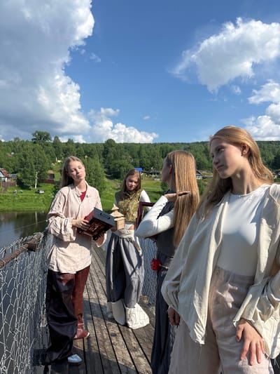 Four young women stand on a wooden suspension bridge on a sunny day. The bridge spans a body of water and is flanked by a chain-link fence. In the background, a lush green forest lines the hillsides, with scattered houses and buildings visible. The sky is a bright blue with fluffy white clouds.
The woman closest to the viewer on the right is wearing a light-colored, loose-fitting outer garment over a white top and light-colored pants. Her blonde hair is down, and she is looking to her right, with her left hand resting on her lap.
To her left, another woman with blonde hair, also down, is wearing a dark long-sleeved shirt and a gray skirt. She is looking towards the woman to her left. A red object is attached to her belt.
In the center, a woman with blonde hair tied back is wearing a vest over a white top and a gray skirt. She is holding a small, intricately designed wooden structure.
On the far left, a woman with blonde hair is wearing a light-colored tunic and dark, wide-legged pants with a patterned print. She is holding a red and black box. She is looking down with her eyes closed.
The scene appears to be set in the city of Gubakha, Russia, given the style of the wooden bridge and the surrounding architecture.