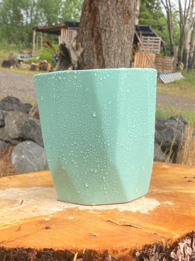 A mint green faceted pot with water droplets is centered on a log in Haines Junction, Canada. The pot is in the foreground, with a blurry background of trees, a wooden structure, and a pile of lumber. Rocks are visible to the left of the pot. The pot is casting a subtle shadow on the log. The overall scene suggests an outdoor, possibly rustic, setting.