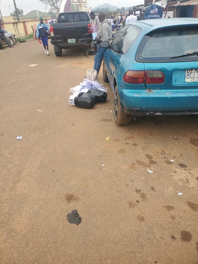An eye-level, medium shot captures a street scene in Ohit, Nigeria. In the foreground, a blue hatchback car is parked on a dirt road. To the right of the car, a pile of white and black plastic bags filled with items lies on the ground. The car's rear bumper is damaged, with red paint showing through. Behind the car, a black pickup truck is also parked.

In the middle ground, a man in a grey hoodie and blue jeans stands near the hatchback, holding a camera. To the left of the pickup truck, a young man in a blue and white tracksuit walks with a red folder. Other individuals are visible in the background, some standing near motorcycles and shops. A "GOLDBERG" sign is visible above some of the shops in the distance. The ground is a mixture of dirt and gravel, with scattered debris including paper and what appear to be oil stains. The sky is overcast.