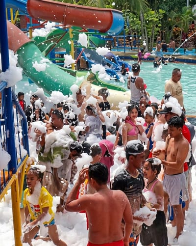 The image depicts a crowded foam party at a water park in Durin Simbelang, Indonesia. Numerous people, primarily children and young adults, are covered in white foam. Many are standing and interacting with each other, while others are closer to the water's edge. Behind the crowd, there are colorful water slides and a swimming pool with other individuals. Lush green trees are visible in the background.