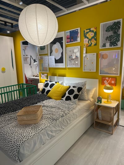 This is a medium shot, eye-level, indoor photograph of a bedroom display. The room has bright yellow walls. A large white spherical paper lantern hangs from the ceiling. The bed is made with white linens and duvet covers featuring a black and white polka dot pattern. Two yellow accent pillows and two black and white polka dot pillows are arranged on the bed. A woven basket sits on the duvet. To the left of the bed is a green crib. On the wall above the bed are several framed pieces of artwork. To the right of the bed is a white nightstand with a yellow lamp, a green clock, and a woven basket on the lower shelf. The flooring is light-colored laminate. The scene appears to be a showroom, possibly at an IKEA store in Siero, Spain, based on a previous interaction. There are no people or animals present in the image.