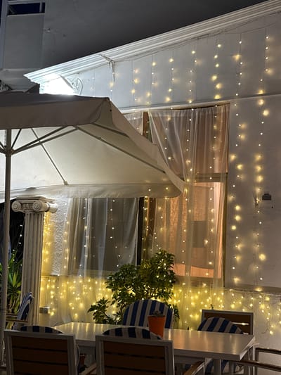 The image depicts an exterior setting, likely a restaurant or outdoor dining area in Altea, Spain, during the evening. Overhead, there's a building structure adorned with numerous strings of small, warm-toned lights, creating a festive ambiance. Semi-transparent curtains or fabric drapes are visible, adding to the decorative layering. A white awning is prominently featured, casting a bright reflection. Part of a white-and-blue striped chair or furniture element is visible towards the upper right corner, contributing to a coastal aesthetic. A decorative pillar with classical design elements also appears in the scene. Green foliage, perhaps from a potted plant, adds a touch of nature to the setting. The overall composition and lighting suggest a pleasant and inviting atmosphere typical of outdoor dining in Altea.