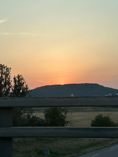 The image captures a sunset over a rolling hill with trees in the foreground, viewed through a barrier. The sky transitions from a pale blue at the top to a vibrant orange and yellow near the horizon, indicating the sun is setting. A dark, tree-covered hill dominates the mid-ground. Two clusters of buildings are visible on the lower slope of the hill. In the foreground, a dark metal barrier runs horizontally across the frame, with two distinct bars. Beyond the barrier, a field with some vegetation and scattered trees is visible. A small, triangular sign is partially visible in the lower left portion of the field. The overall scene suggests a landscape view from an elevated position, possibly a bridge or overpass, in the vicinity of Dommartin-lès-Toul, France.