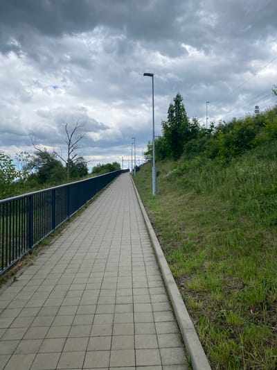 The image shows a paved pedestrian walkway with a black metal railing on the left, bordered by a grassy slope on the right. Several streetlights are visible along the walkway, which curves upwards into the distance. In the background, there are trees, some vegetation, and overhead power lines. The sky is overcast with dark, dramatic clouds. This scene is located in Lahošť, Czechia.