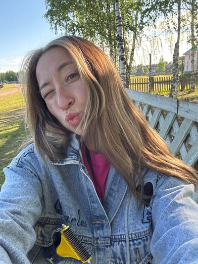 A female subject is depicted in a close-up outdoor selfie, winking her left eye and pursing her lips in a "kissing" expression. She has long, light brown hair with lighter highlights, parted to the side, and visible freckles on her face. She is wearing a light blue denim jacket over a hot pink garment. The jacket features "Charles" embroidered in dark thread on the viewer's left chest area and a dark, possibly black, letter "M" or symbol with a heart outline on the viewer's right chest area. A yellow comb with multiple metal teeth is clipped to the front of her jacket. The scene is set in Ust'-Silayka, Russia, during bright daylight hours, with sunlight illuminating the subject from the right. The background features a dirt path and green grass to the subject's left, with distant trees and utility poles. To the subject's right, a weathered light green wooden lattice fence runs horizontally, behind which stand several birch trees with white bark. Further in the distance, a light pink or peach-colored house with white trim and other structures are visible, indicating a rural or village environment.