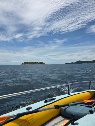 The image shows a view from a boat in the sea, with an island in the distance. The sky is blue with scattered white clouds. The water is dark blue and appears choppy. In the foreground, part of an inflatable boat, possibly a kayak, is visible, along with a paddle. The scene suggests a recreational activity at sea. The location is identified as Bang Sare, Thailand.