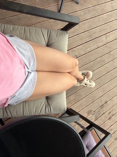 The image shows a person's legs from the knees down, wearing a pink t-shirt and grey shorts. They are sitting on a cushioned chair, which is placed on a wooden deck. The person is also wearing cream-colored Crocs with charms. To the left, a portion of a black table is visible. The scene appears to be outdoors, possibly on a patio or balcony, with wooden planks making up the floor. The location is identified as Семилуки, Russia.