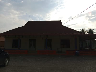 The image shows a low-angle exterior shot of a building with a red corrugated metal roof, located in Cotpuuk, Indonesia. The building has a covered porch area with orange decorative fencing along the bottom. Several windows and doors are visible on the building's facade. To the left, the front portion of a dark-colored car is parked on a paved surface. The sky above is partly cloudy with the sun visible, suggesting it is daytime. Palm trees are visible in the background to the right. The overall scene appears to be a residential or community building in a tropical or subtropical environment.