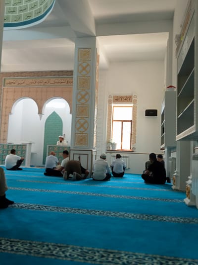 The image depicts the interior of a mosque with a bright blue carpet. Several men are seated on the carpet, facing different directions, some in prayer postures. In the background, a man in traditional attire stands near a green patterned wall. The architecture features decorative elements on columns and walls, with Islamic calligraphy visible. Natural light streams in from a large window. The overall impression is one of a peaceful gathering for prayer. This mosque is located in the city of Besagash, Kazakhstan.