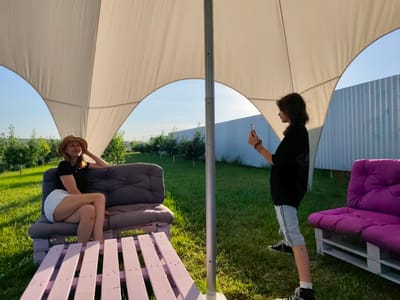 In Nechaiane, Ukraine, two individuals are observed in an outdoor seating area. A woman wearing a straw hat, black t-shirt, and white shorts is seated on a grey sofa. To her right, a second individual, dressed in a black top and light-colored shorts, stands and holds a smartphone, appearing to take a photo. A purple sofa is situated to the right of the standing individual. The scene is partially covered by a large, light-colored canopy supported by a metal pole. Surrounding the seating area is a grassy lawn, with trees visible in the background and a corrugated metal fence. The lighting suggests it is daytime, likely late afternoon, with a clear sky.