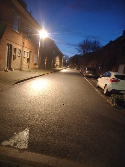 A street view at dusk. A row of parked cars lines the upper-left side of the street. Houses are adjacent to the cars, with lit windows. Streetlights illuminate the scene. The street surface appears wet and reflective. The sky is a mix of blue and dark shades, suggesting twilight. Bare tree branches are visible in the background against the sky.
