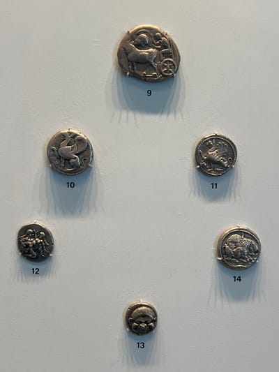 Six ancient silver coins, numbered 9 through 14, are displayed against a plain, light gray vertical surface, secured by small, transparent clips. Shadows indicate an overhead light source.
Coin 9, located at the top center, is an irregularly shaped silver piece depicting a bull or ox pulling a two-wheeled chariot, with a figure seated within the cart.
Coin 10, positioned mid-left, is a circular silver coin featuring a winged griffin or mythical creature facing left.
Coin 11, at mid-right, is a circular silver coin showing a standing winged lion with a raised paw, facing left.
Coin 12, situated at the bottom-left, is a circular silver coin illustrating two figures, possibly human, in an interactive or combative pose.
Coin 13, centered at the bottom, is a circular silver coin presenting a frontal, stylized mask-like face.
Coin 14, at the bottom-right, is a circular silver coin depicting a scene of two animals, likely a lion attacking another large animal such as a bull.
This exhibit is located within the Harvard Art Museums in Cambridge, Massachusetts, United States.