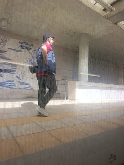 The media file shows a person from the side, walking on a tiled platform. The person is wearing a dark baseball cap, glasses, a bright pink t-shirt, a blue puffer vest, and dark pants. They are also carrying a dark backpack. The platform appears to be outdoors, possibly at a train or bus station, with a concrete ceiling overhead and a tiled floor. There is a patterned mural on the wall behind the person. The lighting suggests it is daytime, with bright sunlight casting shadows. The overall scene seems to be a casual, everyday moment. There is no visible text in the image.