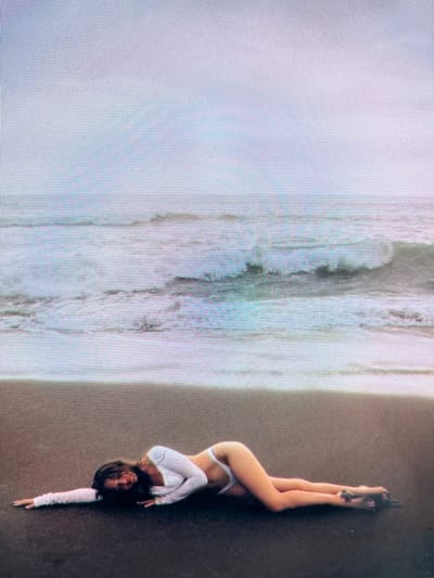 The image shows a woman lying on a dark sandy beach with the ocean in the background. She is wearing a white long-sleeved top and white bikini bottoms. The waves are crashing on the shore. The sky is overcast. The image does not contain any information about the city of Navahrudak, Belarus.