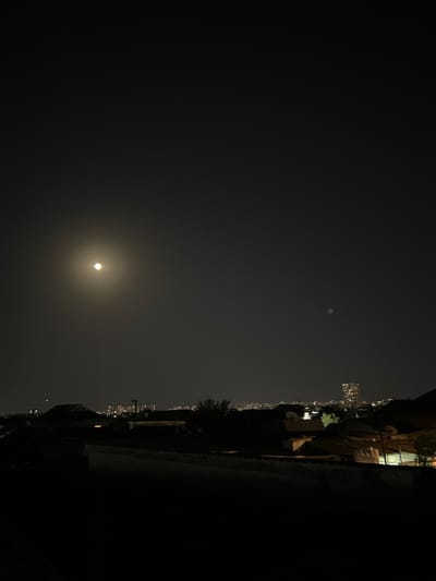 The image captures a nighttime scene of a city skyline, possibly Sabunchu, Azerbaijan, under a bright moon. The foreground is dominated by dark rooftops and silhouettes of buildings, suggesting a residential or urban area. In the mid-ground, various structures, including houses with visible roofs and some with illuminated windows, are discernible. A prominent tall building with many lit windows stands out in the distance, indicating a more developed part of the city. The sky is dark with the moon prominently featured on the left side, surrounded by a faint halo of light. A smaller, faint light source or object is visible in the sky to the right of the moon. The overall impression is a quiet, nocturnal urban landscape.