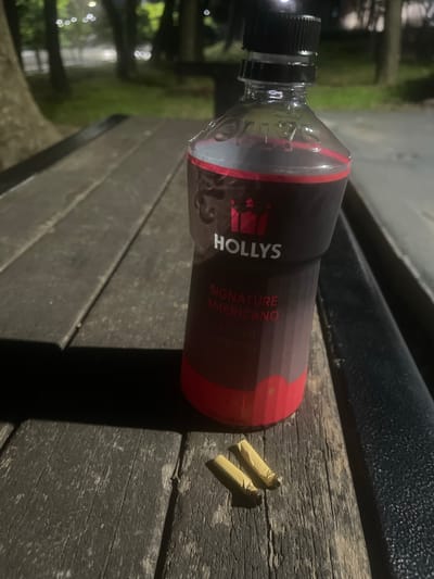 A bottle of HOLLYS Signature Americano is placed on a wooden picnic table outdoors, with two cigarette butts resting on the table beside it. The bottle has a black cap and a dark label with red accents, featuring the "HOLLYS" logo in white. Text on the bottle reads "SIGNATURE AMERICANO" in English and what appears to be Korean script below it. The scene suggests an evening setting, as the background is dark with some blurred lights that could be streetlights or building lights. The wooden table shows signs of wear, indicating it is likely in a park or public outdoor area. The presence of the cigarette butts suggests someone has recently been smoking. The overall mood is one of quiet solitude or a brief moment of rest.
