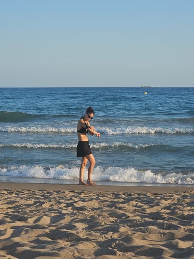 A woman is performing a fitness routine on a sandy beach in Castelldefels, Spain. She is wearing a black bikini top and black shorts, and sunglasses. The waves are gently lapping at the shore, and the sky is clear and blue, indicating a sunny day. The sand is imprinted with footprints, suggesting recent activity on the beach. The scene appears peaceful and idyllic, with the woman engaged in her exercise. The time of day is likely afternoon, given the angle of the sunlight casting shadows on the sand.