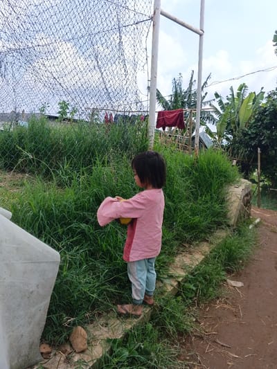 This image depicts an outdoor scene in Maw Hit, Myanmar, under a partly cloudy sky. In the midground, a young child, appearing to be wearing a pink and red long-sleeved shirt, light blue trousers, and dark flip-flops, stands in profile, facing toward the right. The child is holding a yellow spherical object with both hands. To the left foreground, a portion of a light-colored, angular concrete structure is visible. The ground immediately around the child consists of dense green grass, separated from a dirt path on the right by a low concrete curb. In the background, a chain-link fence, partially obscured by additional netting, spans horizontally. Two white metal posts support this fence, one forming an inverted U-shape. Beyond the fence, various green plants, including several large-leafed banana trees, are visible. Multiple pieces of clothing, including a prominent red garment, are hanging to dry on a structure behind the fence.
