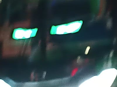 A blurred, dark image features two bright, white, rectangular shapes outlined in green, resembling glowing eyes. Below, faint red and white shapes suggest possible reflections or obscured details. The background is dark and indistinct. The overall impression is of a shadowy figure with luminous eyes.
