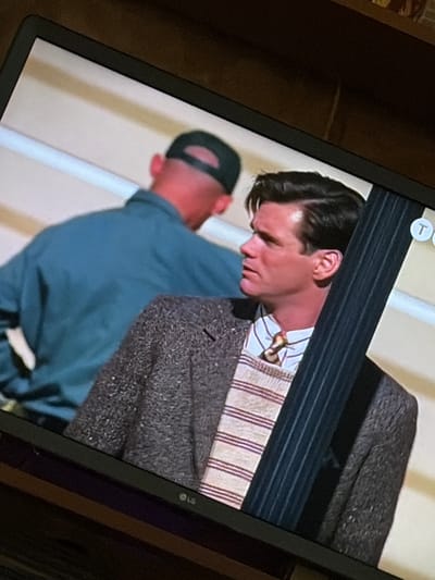 The image is a photograph taken from an angle, likely of a television screen displaying a scene. In the foreground, a man with dark, slicked-back hair and a light complexion is visible from the chest up, looking to his left. He is wearing a tweed jacket over a striped sweater and a tie. Behind him, another person wearing a dark baseball cap and a teal jacket is seen from the back. The background consists of a light-colored wall or structure with horizontal lines. The scene appears to be indoors, possibly on a set or a street with architectural elements. There is no indication of the location being Osolynka, Ukraine, or any other specific geographical place.
